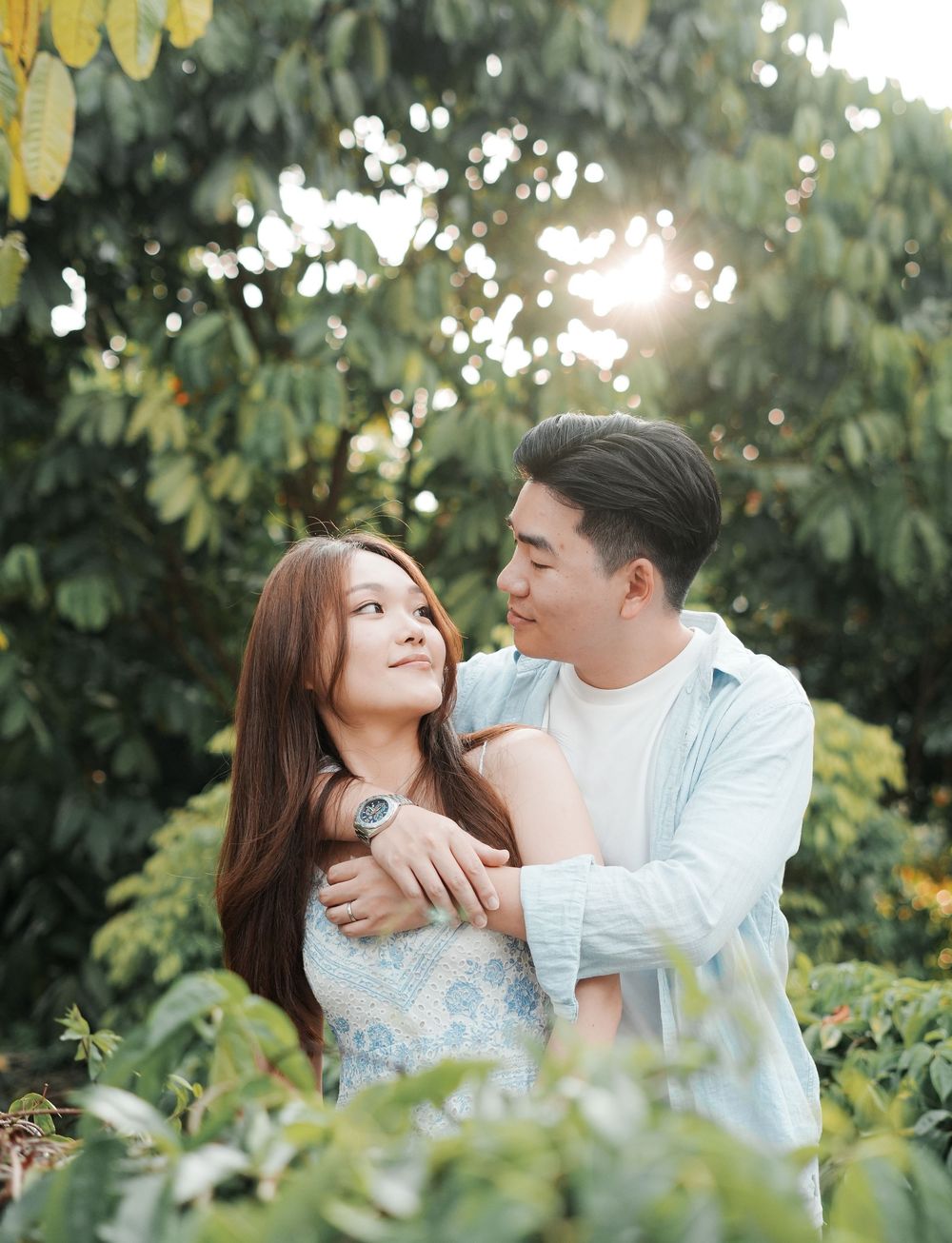Couple embracing in a lush green garden with sunlight filtering through trees, sharing a romantic moment outdoors.