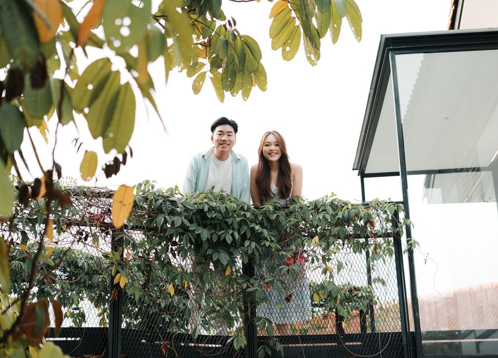 A smiling couple standing on a lush green balcony surrounded by plants and foliage.