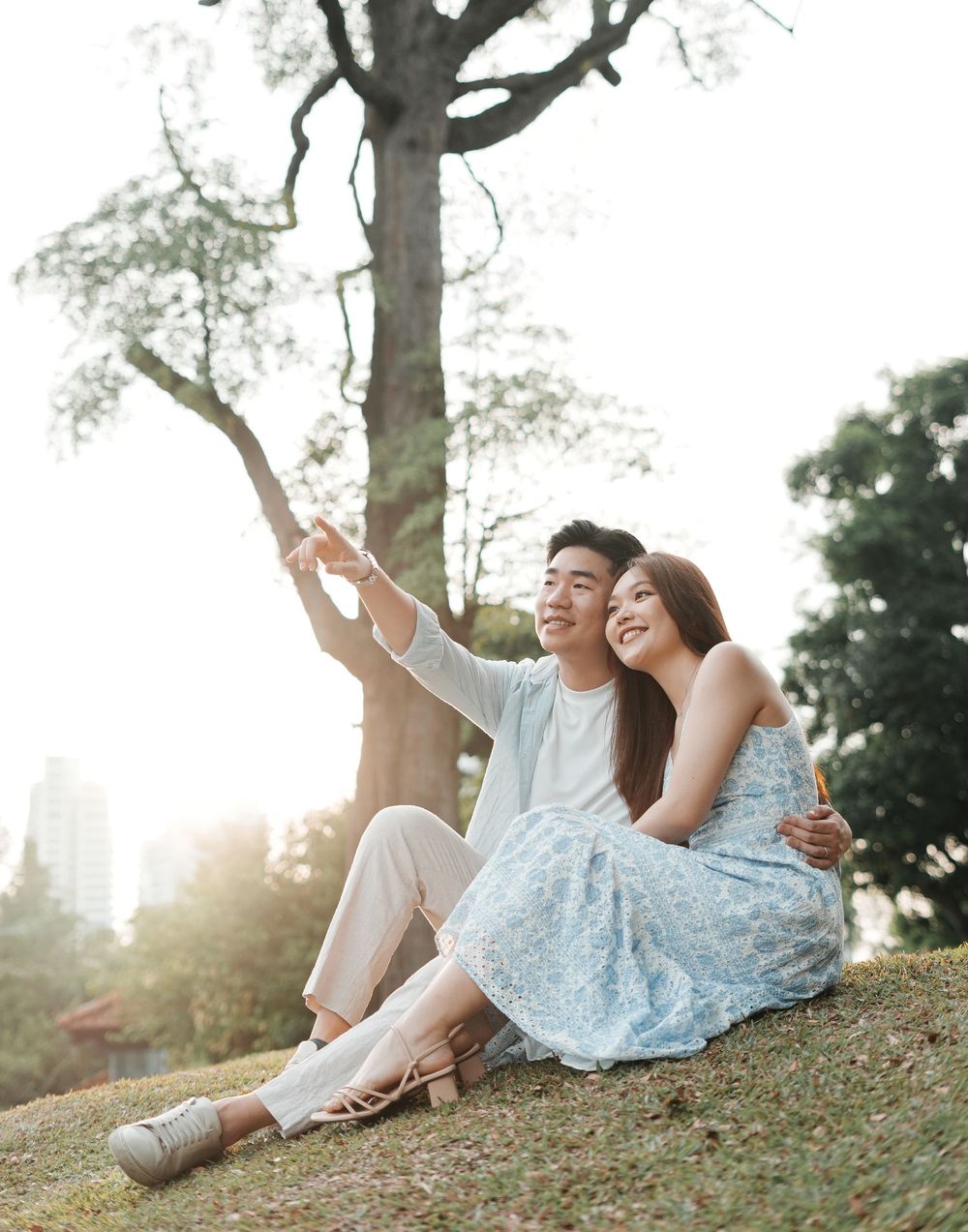 Asian couple sitting on grassy hill under large tree, man pointing while woman smiles.