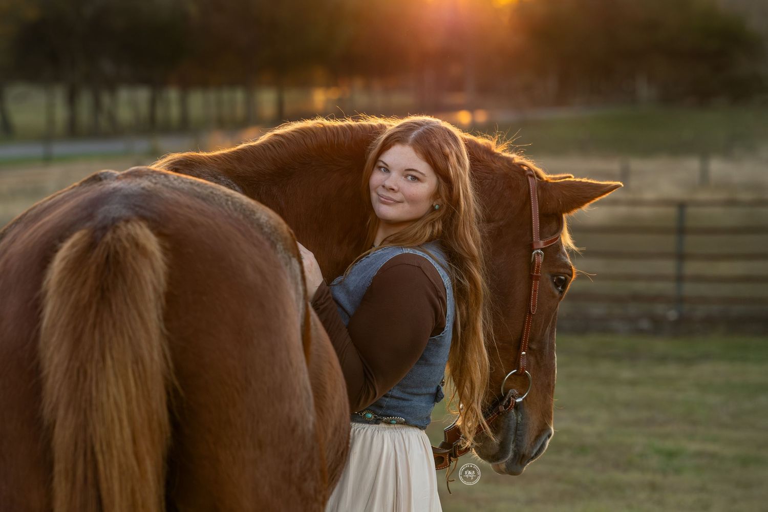 Young woman with long red hair smiling beside a chestnut horse at golden sunset on a farm.