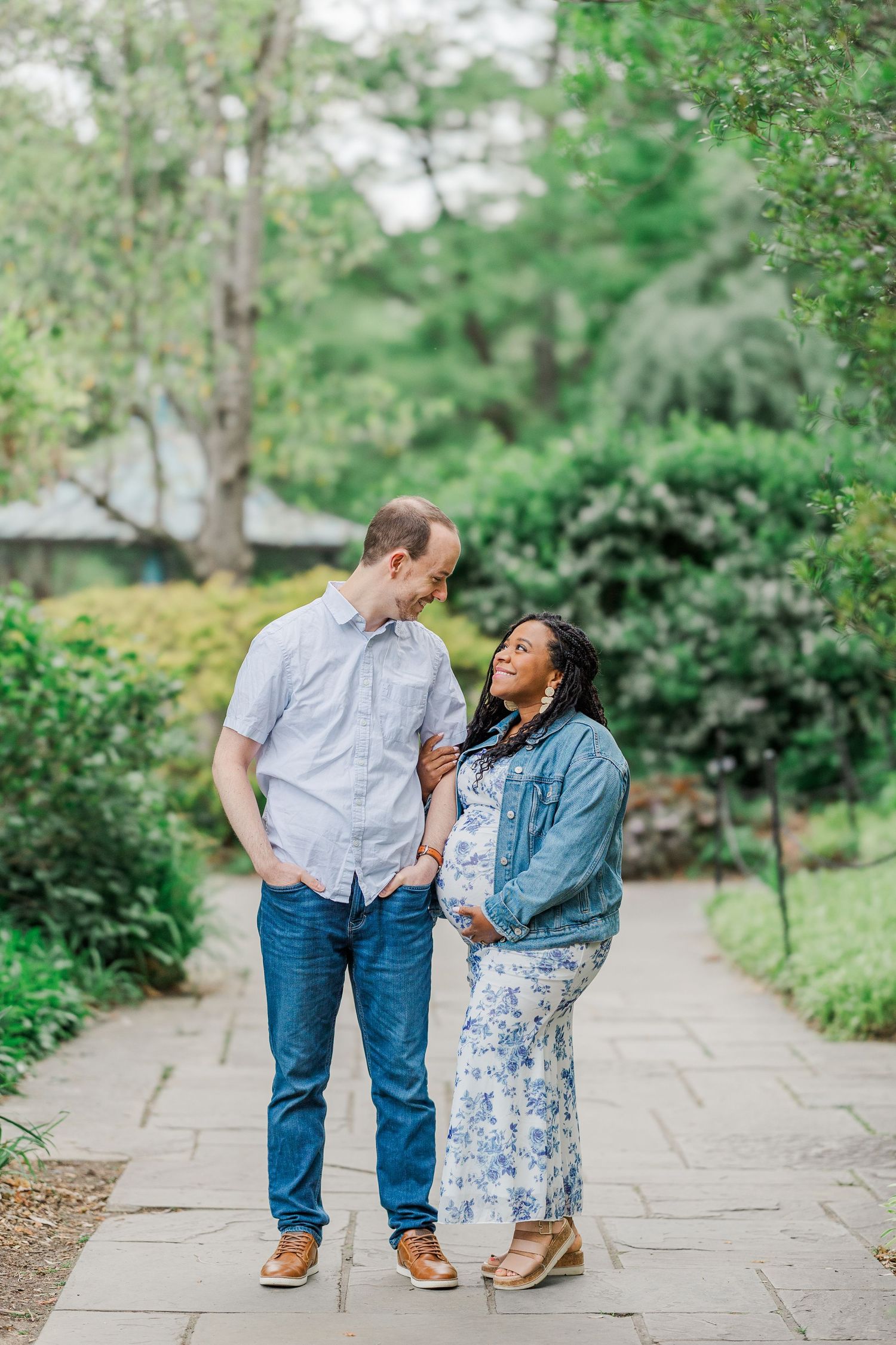 Expecting couple stands on garden path, man in blue shirt and jeans, woman in floral dress and denim jacket cradling baby bump.