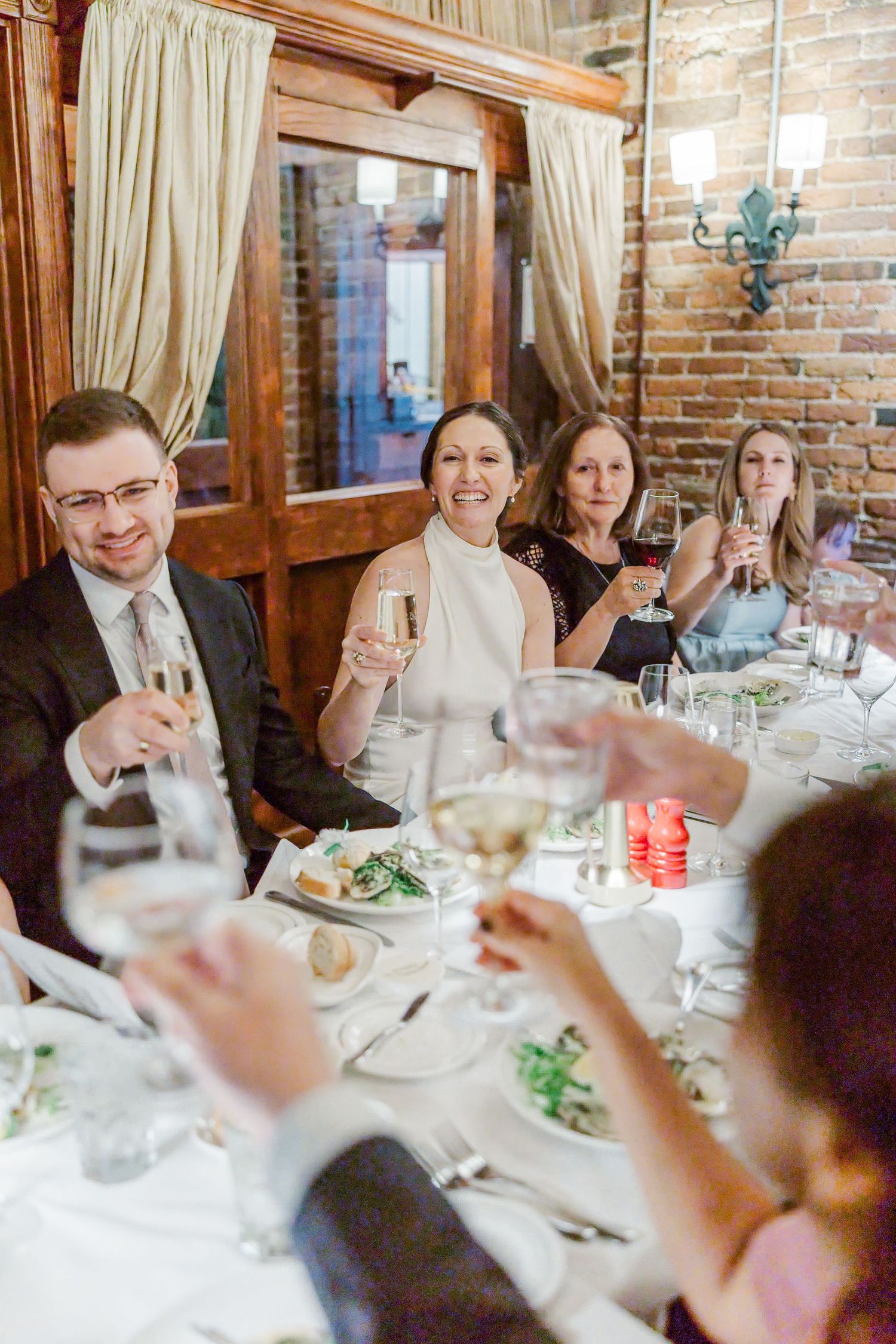 Wedding guests raising glasses in a toast at an elegant dinner reception with brick walls and candlelit ambiance.