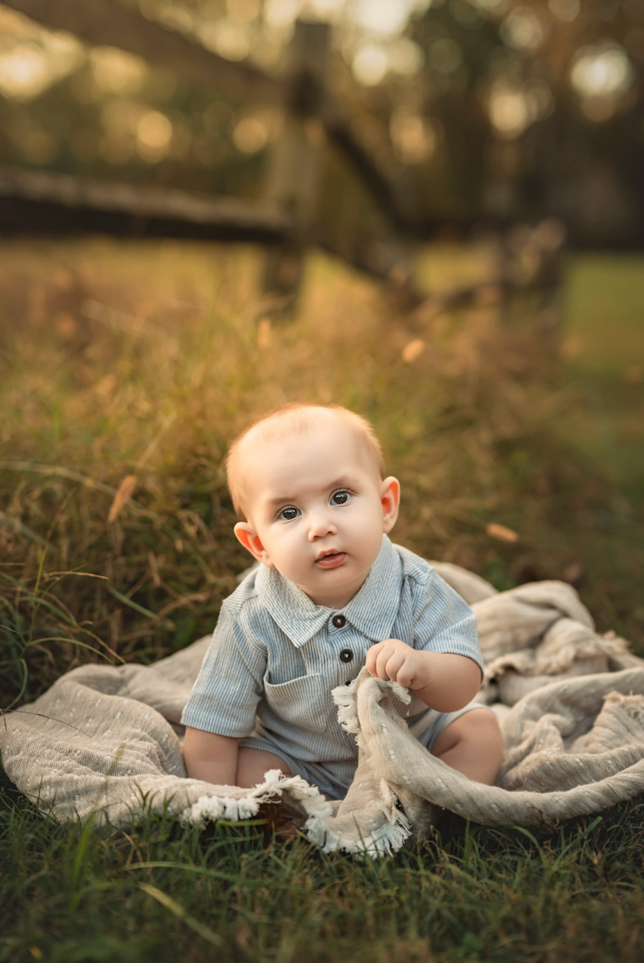 Adorable baby in blue romper sitting on a blanket outdoors in golden hour sunlight.