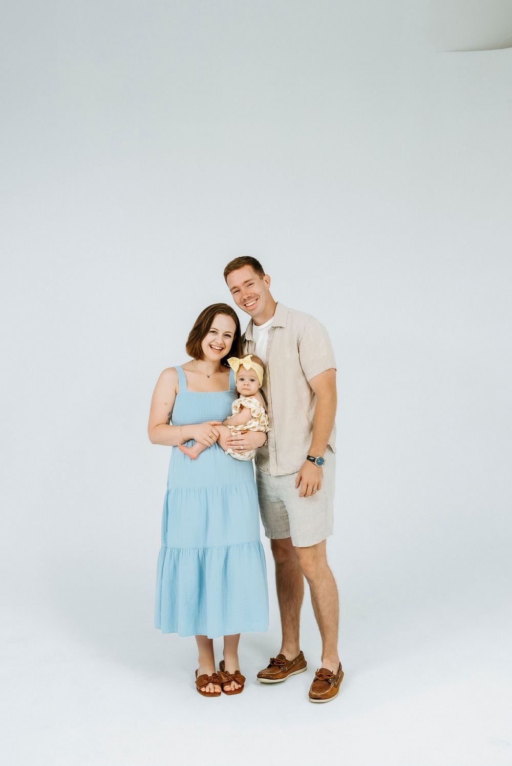 A smiling family of three poses in a bright studio, with mom in a blue dress and dad holding their baby girl.