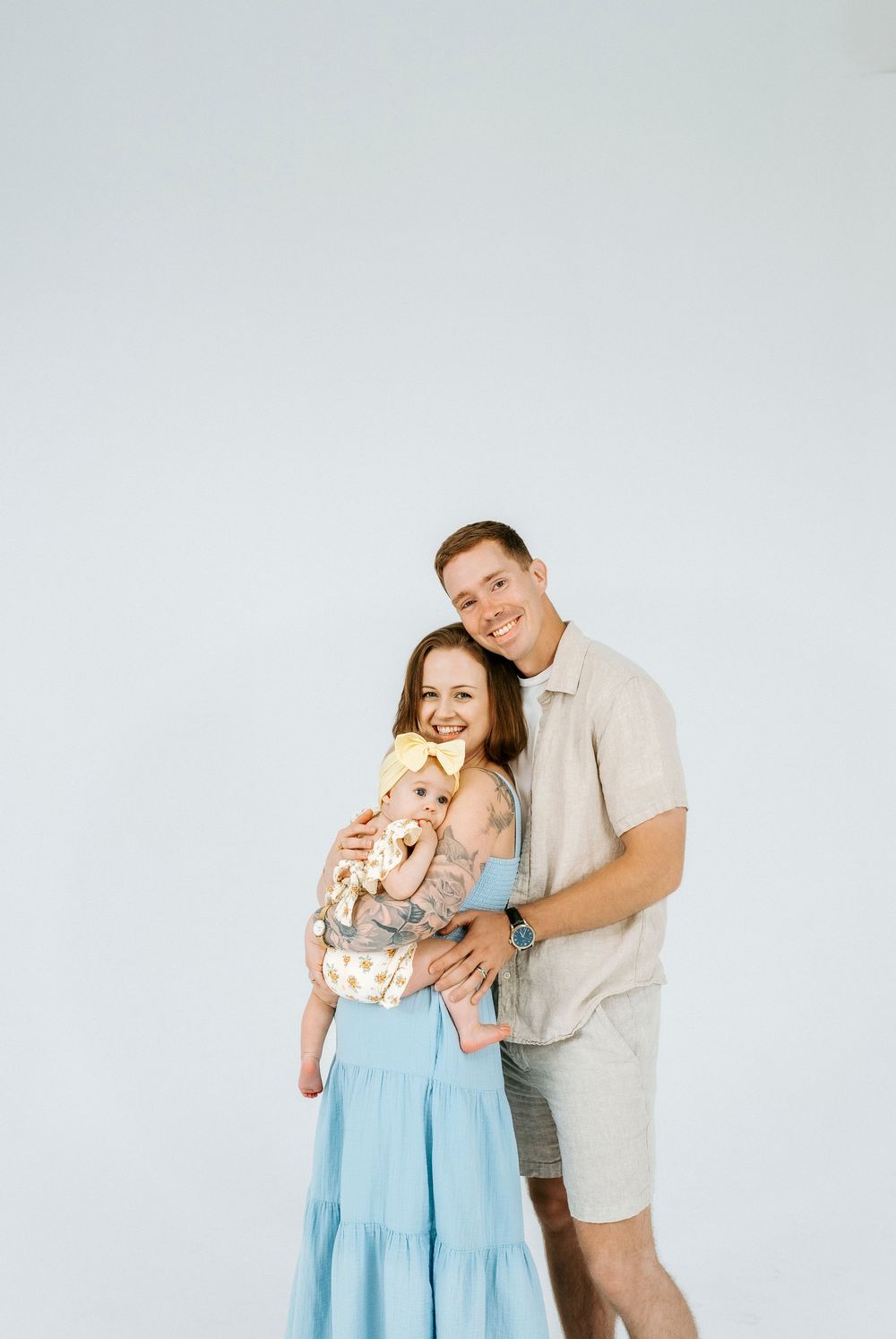 Smiling couple in a photo studio, man embracing woman in blue dress holding a baby in a carrier.