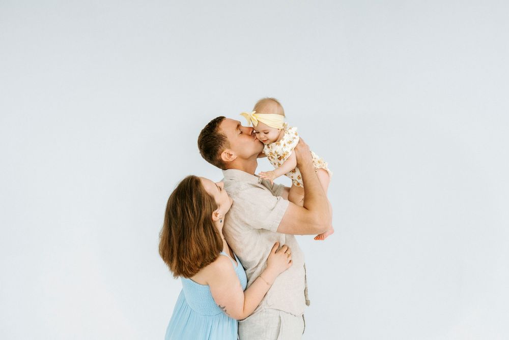 Happy young couple kissing their smiling baby against a light gray studio background.