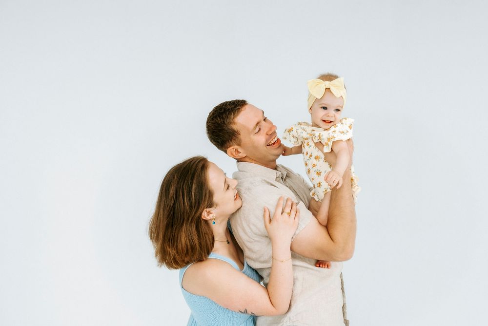 Happy young family with mother, father, and baby girl in floral outfit posing together on white background.