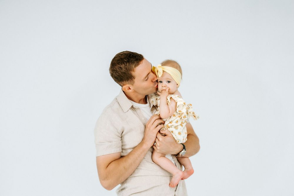 A loving father kisses his baby girl in a floral outfit and yellow bow headband against a white background.