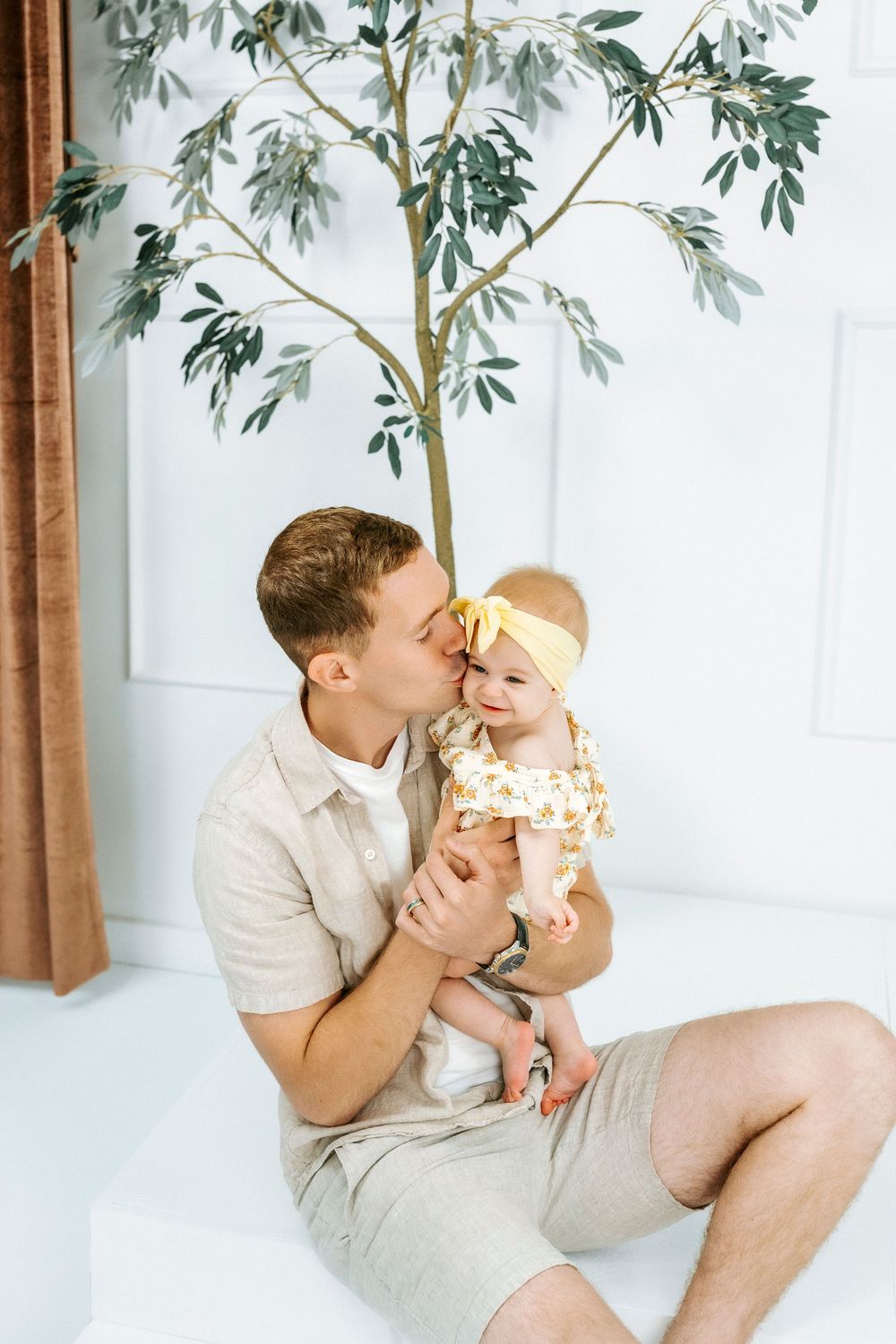 A father kisses his smiling baby girl in a yellow bow and floral outfit while sitting indoors near a decorative tree.