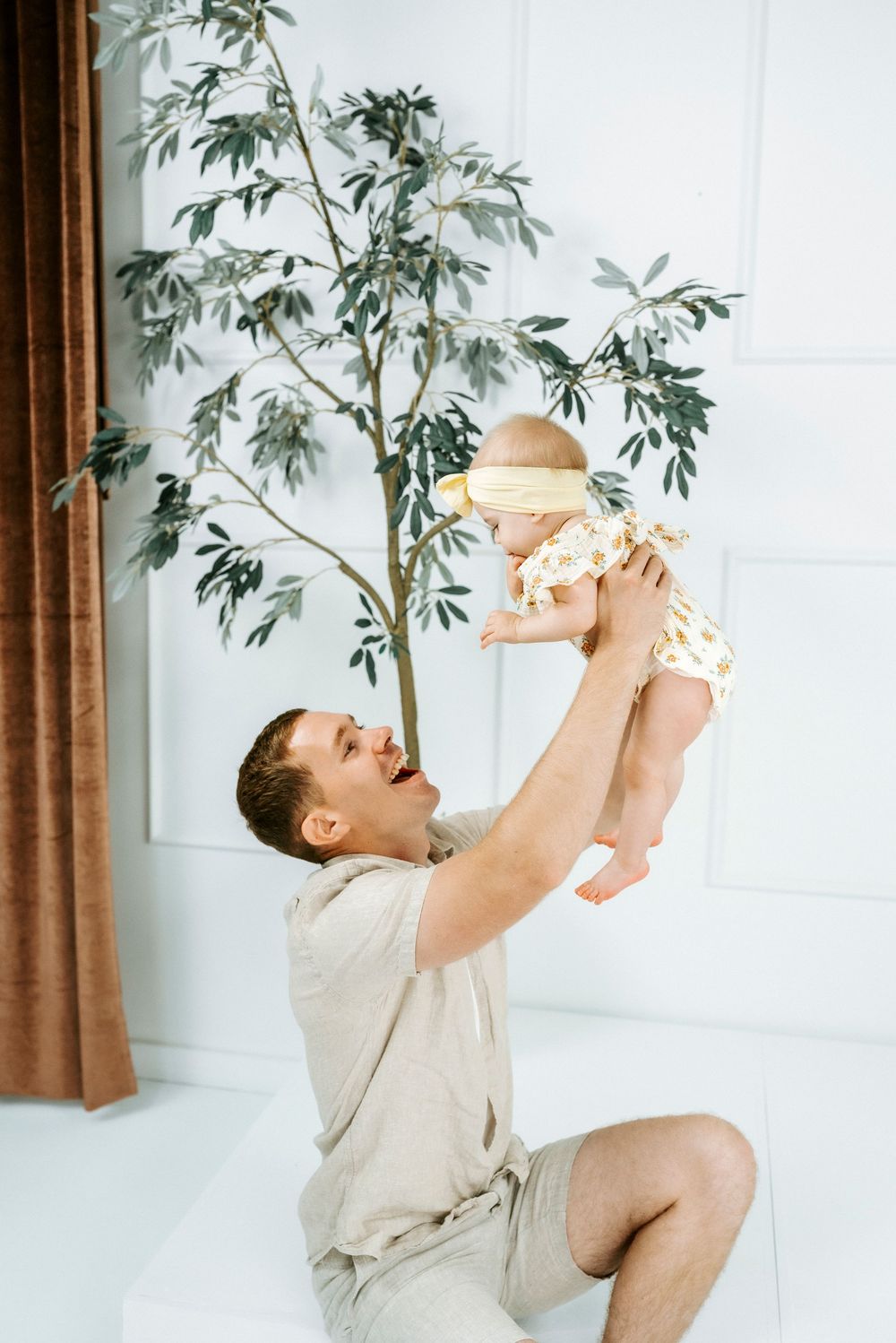 A joyful father lifts his smiling baby overhead in a bright white room with a decorative green tree.