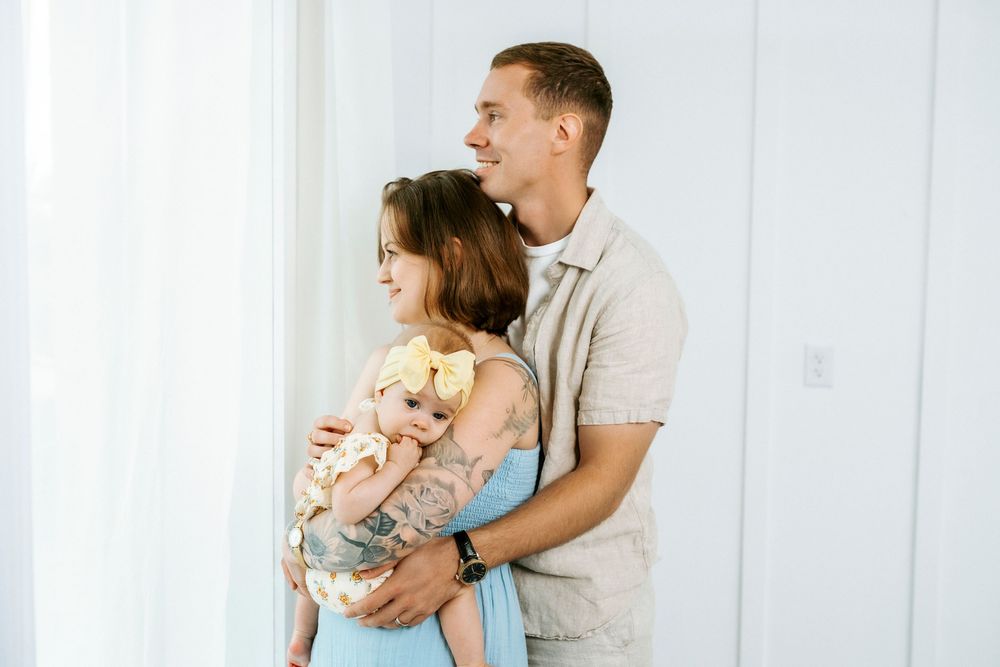 A young couple smiles warmly, man embracing woman holding a teddy bear, standing by a bright white curtained window.