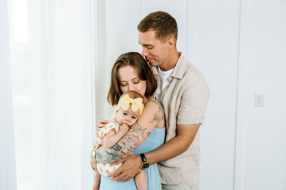 A couple tenderly holds their newborn baby wrapped in a yellow bow swaddle near a bright white curtain.