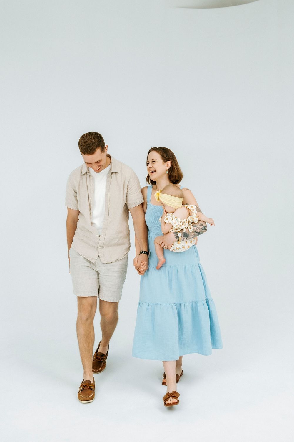 A smiling couple walks together, woman in blue dress holding flowers, man in beige shorts and shirt, on white background.