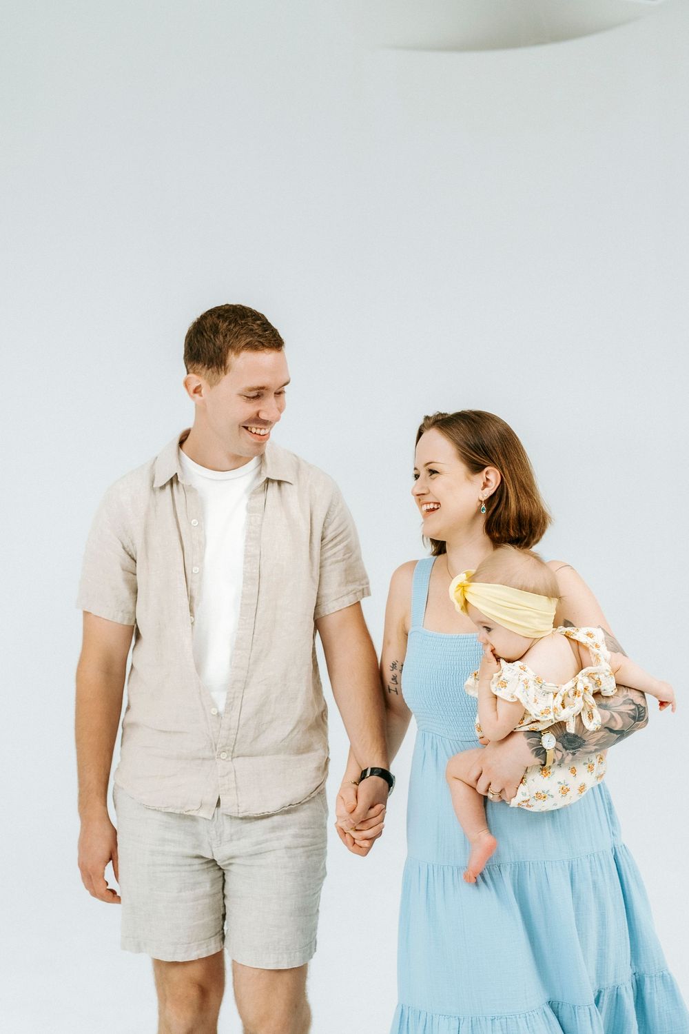Happy young couple holding hands and smiling, with woman in blue dress cradling a baby in yellow outfit.