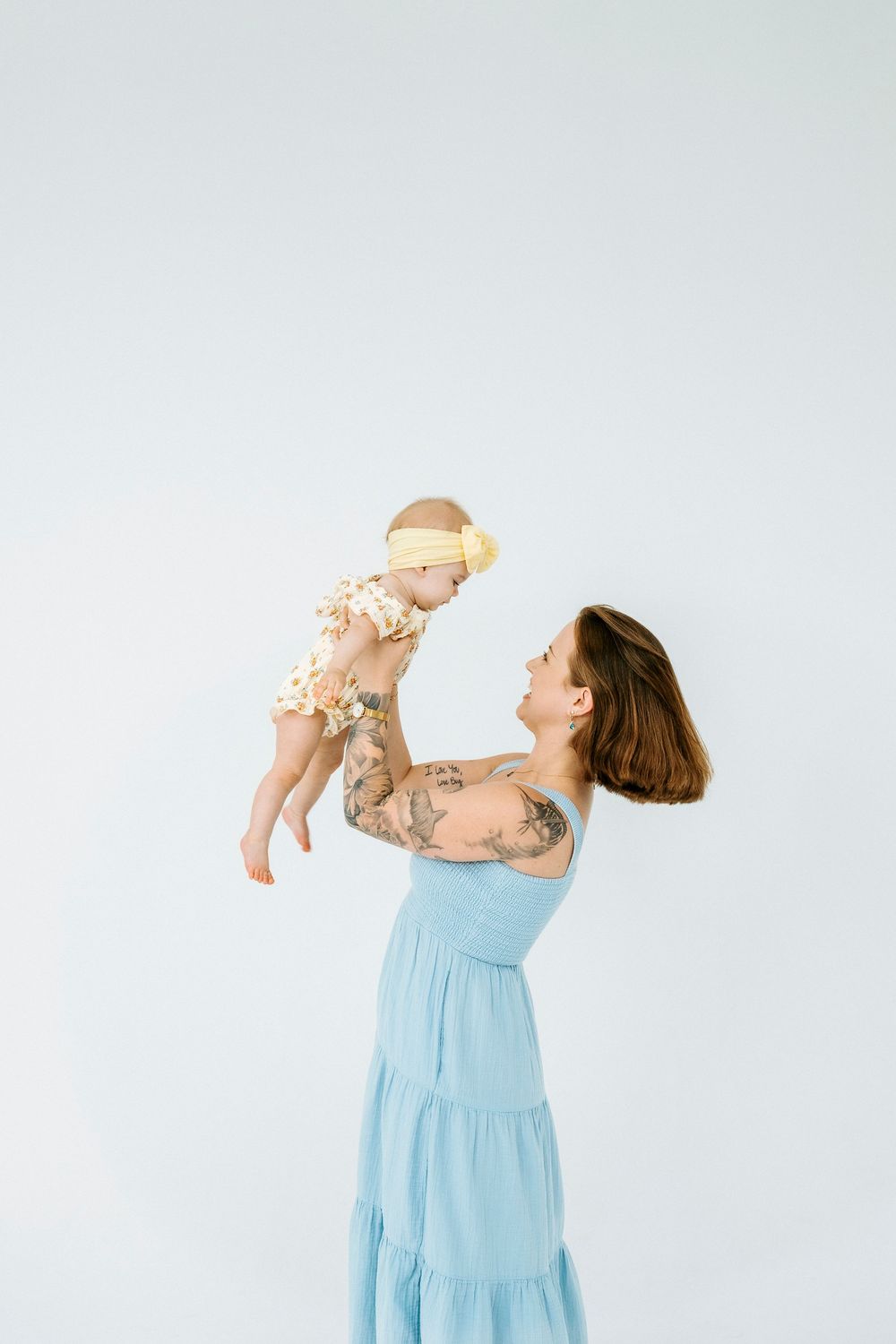 Tattooed mother in blue dress lifting smiling baby wearing floral outfit and yellow headband against white background.