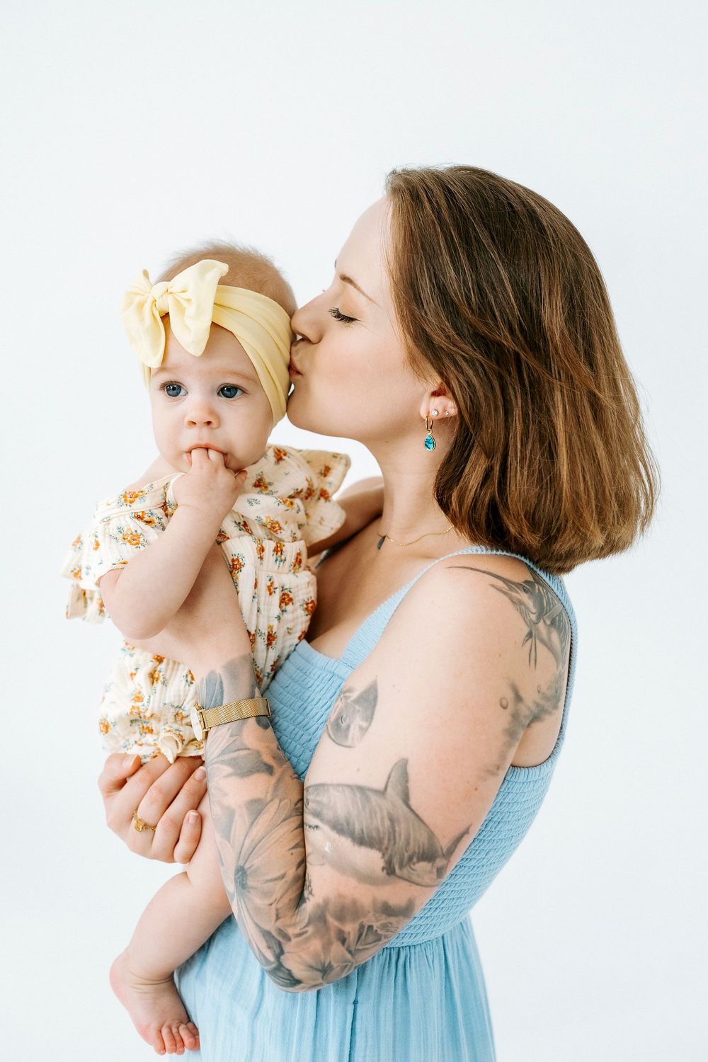 A tattooed mother in a blue dress kisses her baby girl wearing a floral outfit and yellow bow headband.