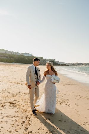 A bride and groom walk hand-in-hand on a sunny beach, she in lace gown with bouquet, he in beige suit.