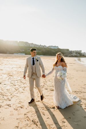 Bride and groom walk hand-in-hand on a sunlit beach, she in lace gown with bouquet, he in beige suit.