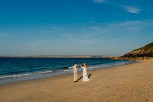 Bride and groom holding hands on a sandy beach with blue ocean and clear sky backdrop.