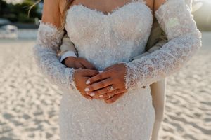 Close-up of bride and groom hands intertwined, showcasing wedding rings on a lace wedding dress at sunset.