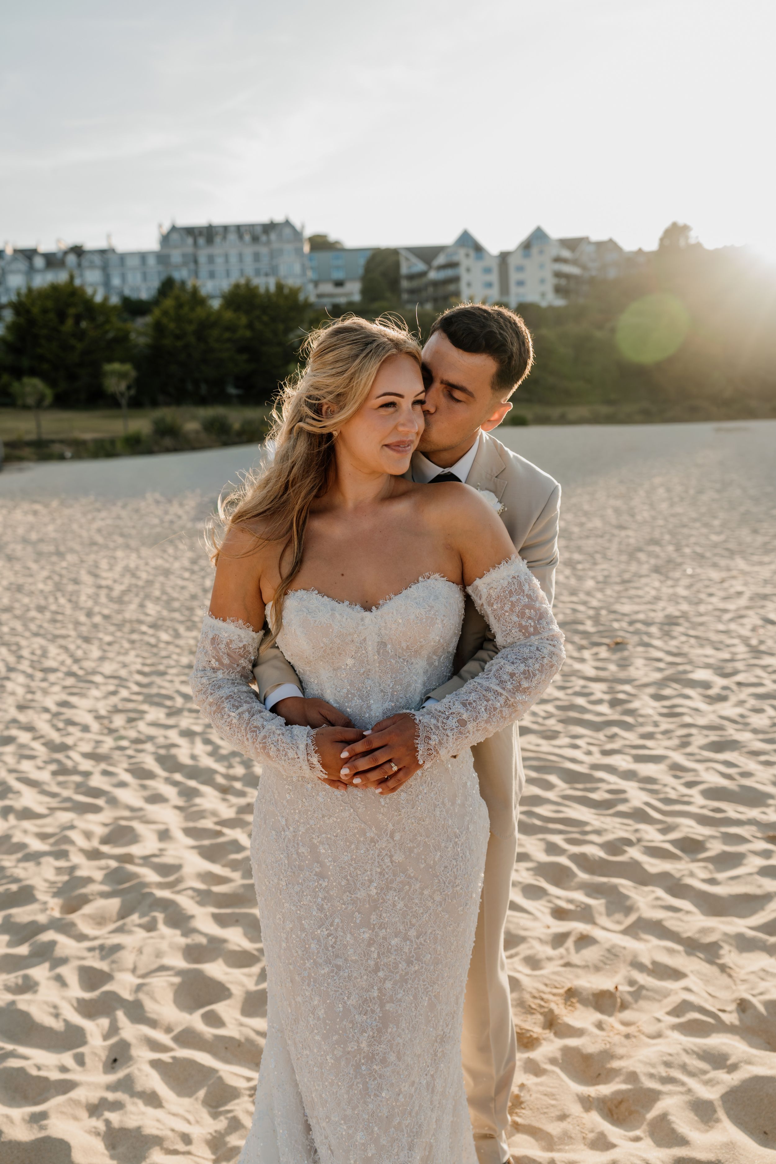 A couple in wedding attire embraces on a sandy beach at sunset, the bride in a lace gown and the groom in a tan suit.
