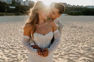 Couple embracing on a sandy beach at golden hour, bride in lace off-shoulder gown, groom kissing her cheek.