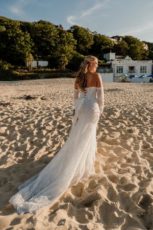 Bride in lace off-shoulder wedding gown with long train stands on sandy beach, trees and white building behind her.
