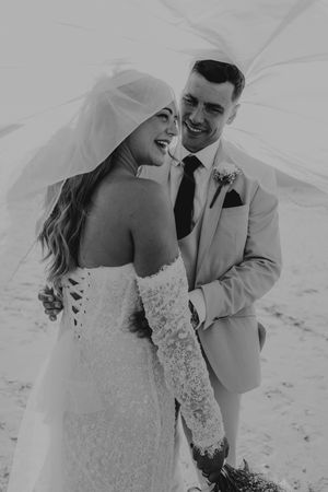 Black and white photo of a smiling bride and groom embracing under a flowing veil on their wedding day.