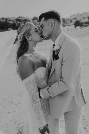 Black and white photo of a bride and groom sharing a romantic kiss on a sandy beach on their wedding day.