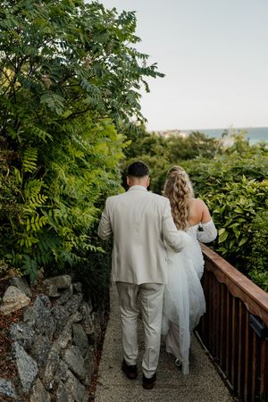 A bride and groom walk hand-in-hand along a wooden railing path surrounded by lush greenery with an ocean view.