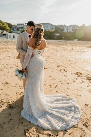 Bride and groom share a romantic moment on a sandy beach, bride wearing elegant lace off-shoulder gown with train.