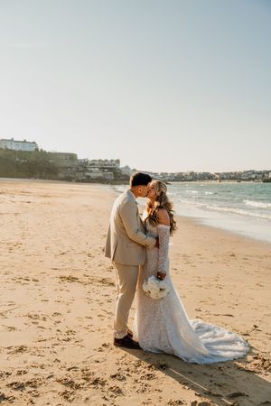 Bride and groom kissing on a sandy beach at golden hour, bride in lace gown holding white bouquet.