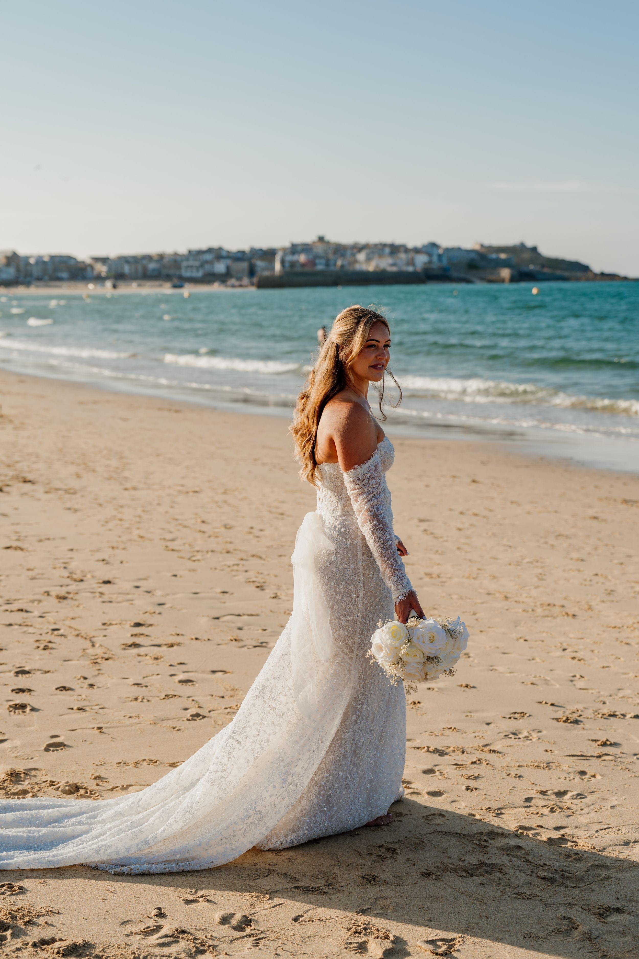 Bride in white lace gown holding bouquet on sandy beach with ocean waves and coastal town in background.