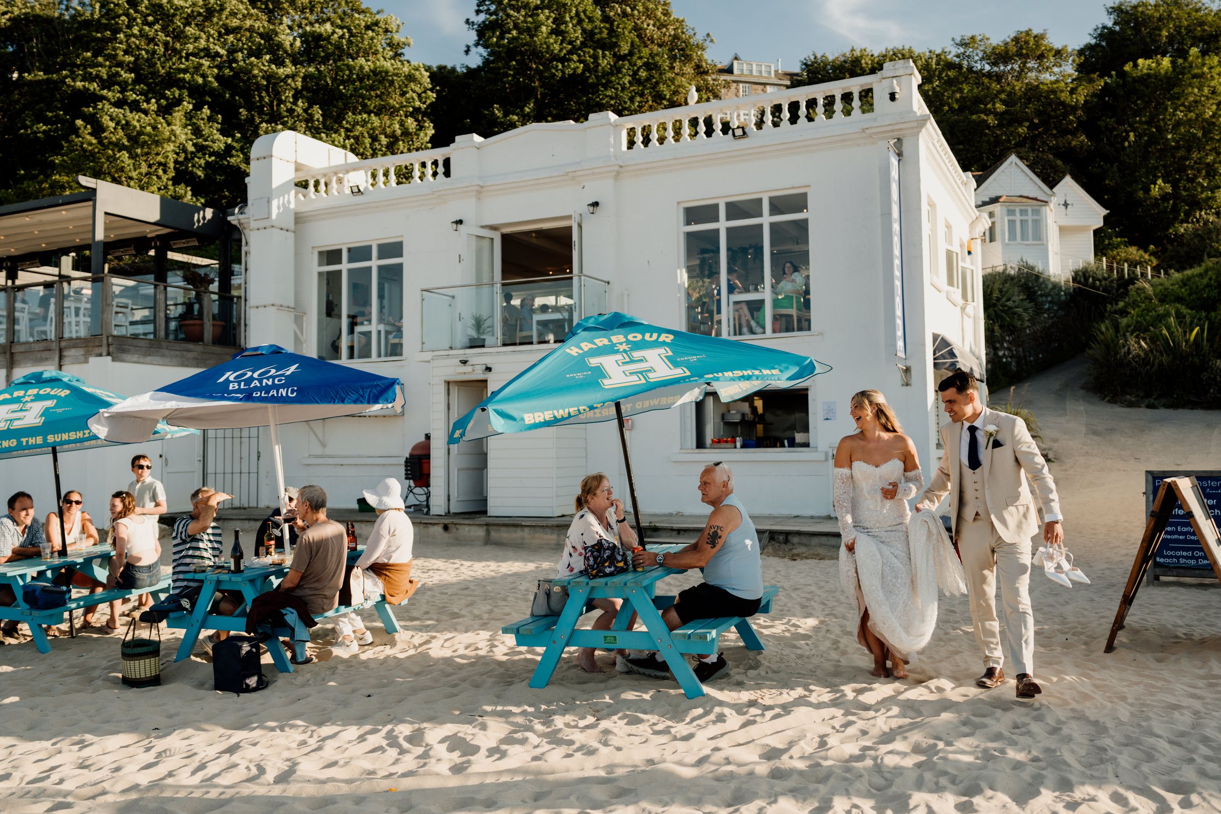 Couple in wedding attire walking on sandy beach near white seaside cafe with blue umbrellas and guests dining outside.