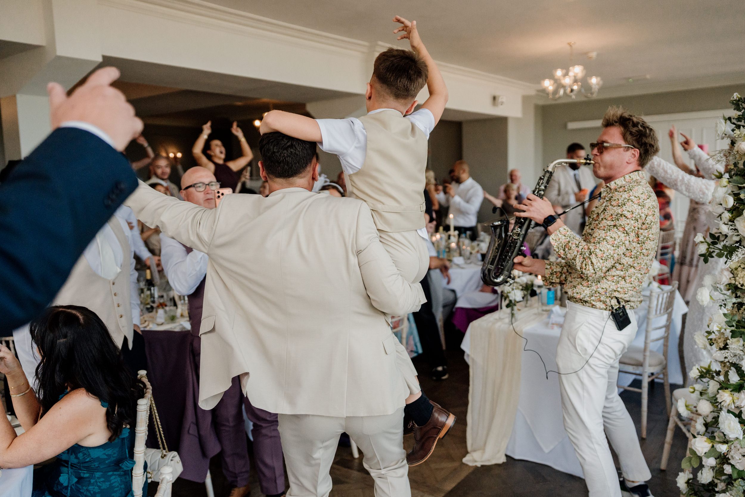 Lively wedding reception with guests dancing, a child lifted in the air, and a saxophonist performing in elegant venue.