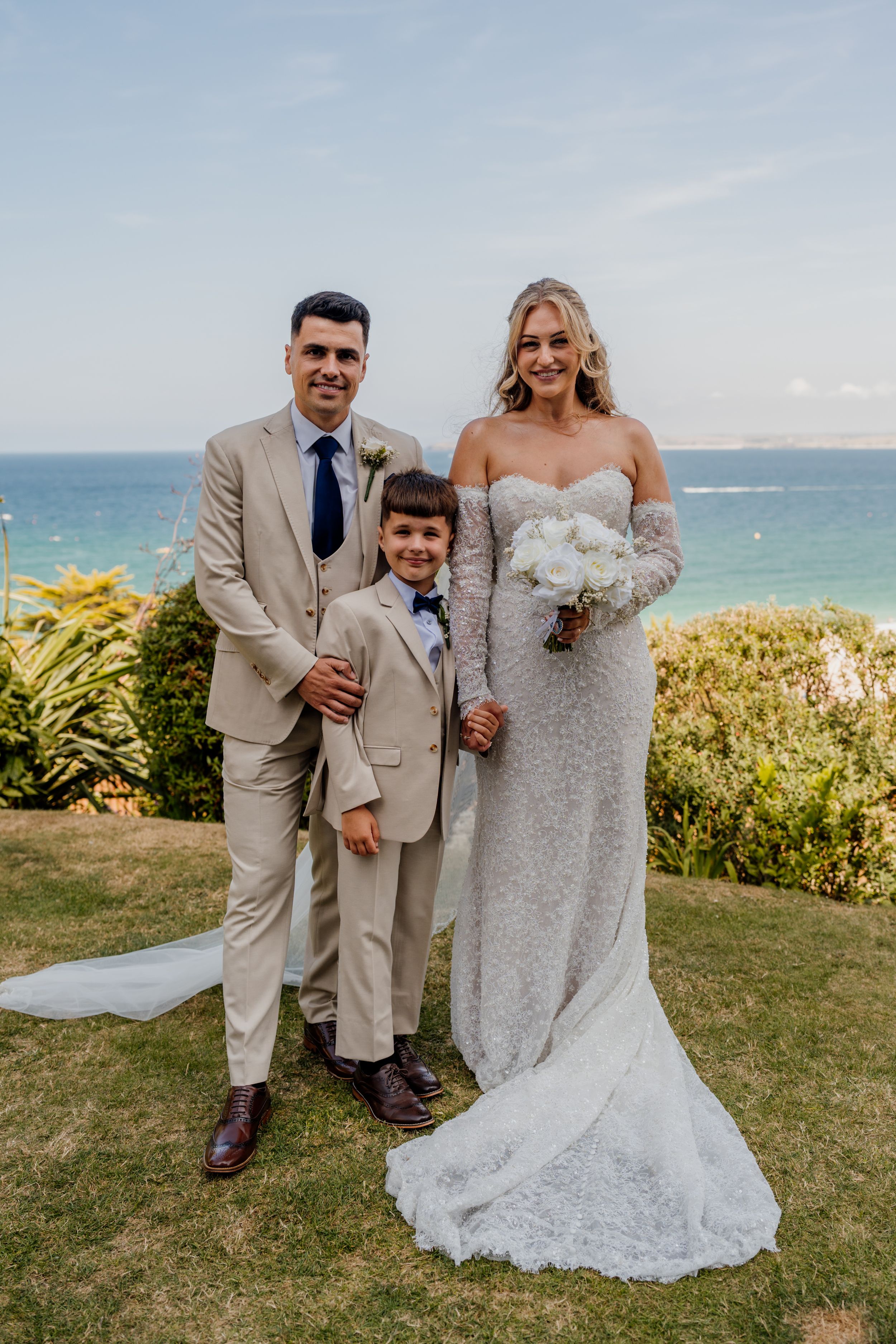 Bride in lace gown holding white bouquet stands with groom in tan suit and young boy at oceanside wedding.