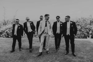 Groom in light suit walks confidently with five groomsmen in dark suits and sunglasses outdoors in black and white photo.