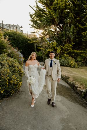 Bride and groom walk hand-in-hand down a garden path, she in a white gown, he in a beige suit.