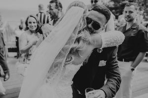 Black and white photo of a groom in sunglasses embracing his bride in a wedding veil at an outdoor reception.