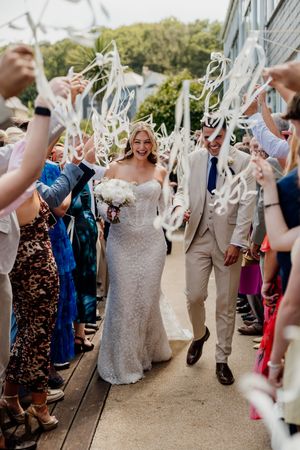 Bride and groom walk through a ribbon wand send-off surrounded by cheering wedding guests outdoors.
