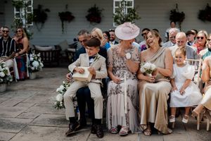 Wedding guests laughing and smiling during outdoor ceremony, including boy in suit, woman in pink hat, and girl in white dress.