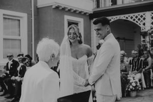 Bride and groom smiling during outdoor wedding ceremony while officiant leads vows before seated guests.