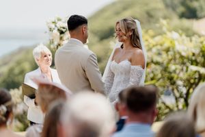 Bride and groom exchange vows outdoors with an officiant, smiling guests seated behind them at a scenic coastal wedding.