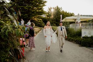 Bride and groom walk hand-in-hand down a sunlit garden path, guests lining the sides at an outdoor wedding.