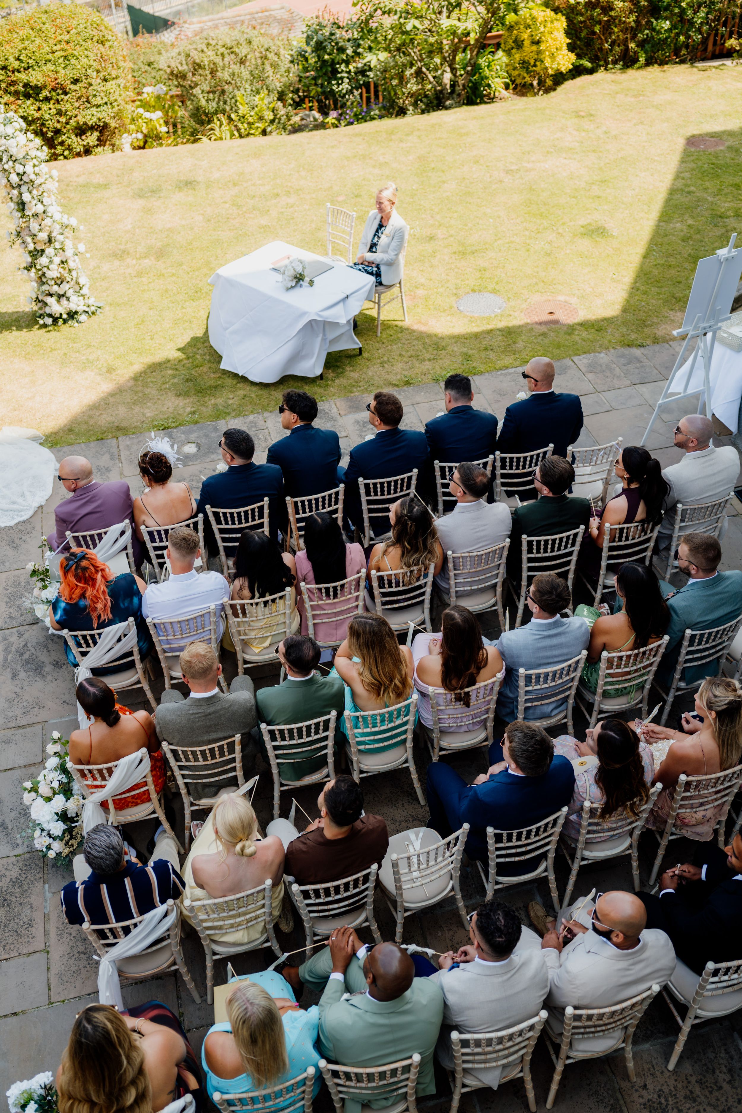 Aerial view of outdoor wedding ceremony with guests seated in rows facing officiant at white draped altar on sunny day.