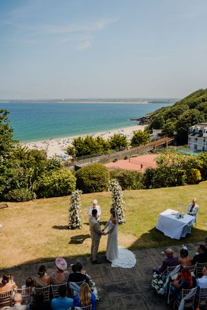 Outdoor wedding ceremony on a lawn overlooking a stunning turquoise beach, with guests seated and couple exchanging vows.