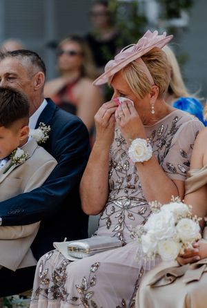 Emotional wedding guest in pink fascinator hat wiping tears during outdoor ceremony, wearing beaded dress with wrist corsage.