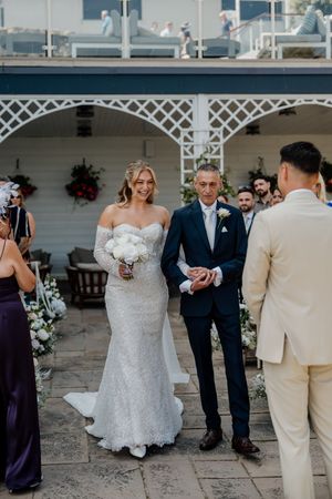 Bride in lace gown walks down aisle with escort toward groom at outdoor wedding ceremony.