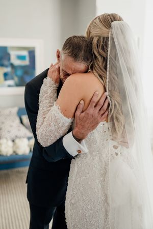 A bride in a beaded gown and veil shares an emotional hug with her father in a suit on her wedding day.