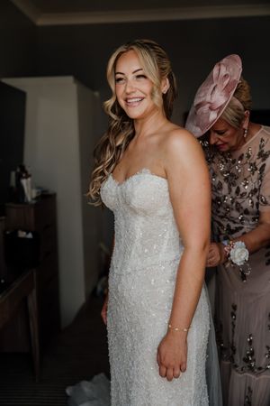 Smiling bride in a strapless beaded wedding gown being helped by a woman in a pink fascinator hat.